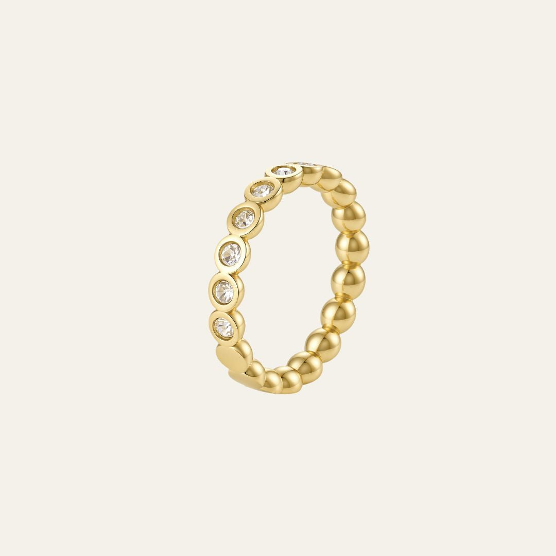 A gold beaded ring with a row of small clear gemstones set along the front, shown on a light cream background.