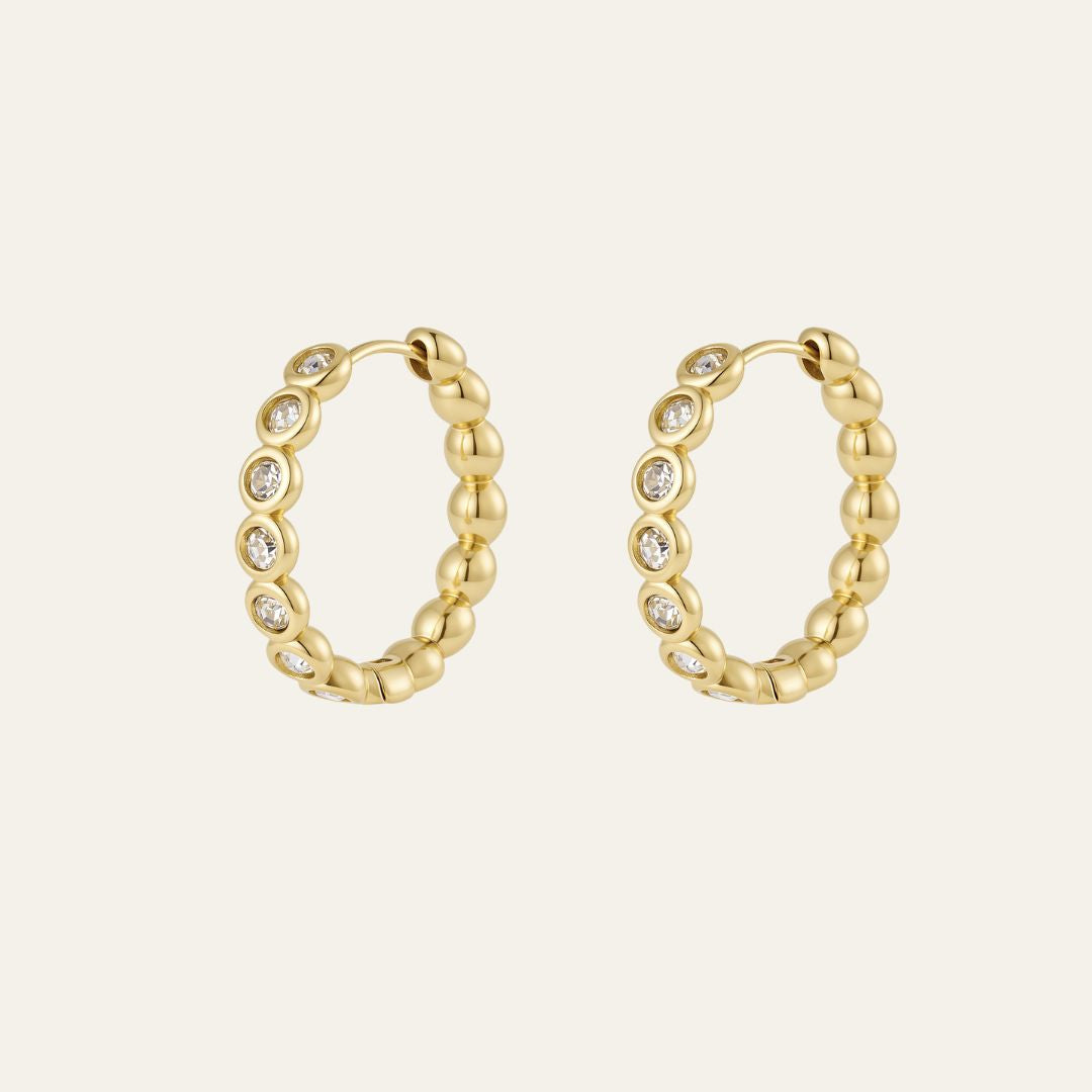 A pair of gold hoop earrings with a textured, bezel design and beaded outer edge. The hoops have small clear stones set along the front, shown on a light cream background.