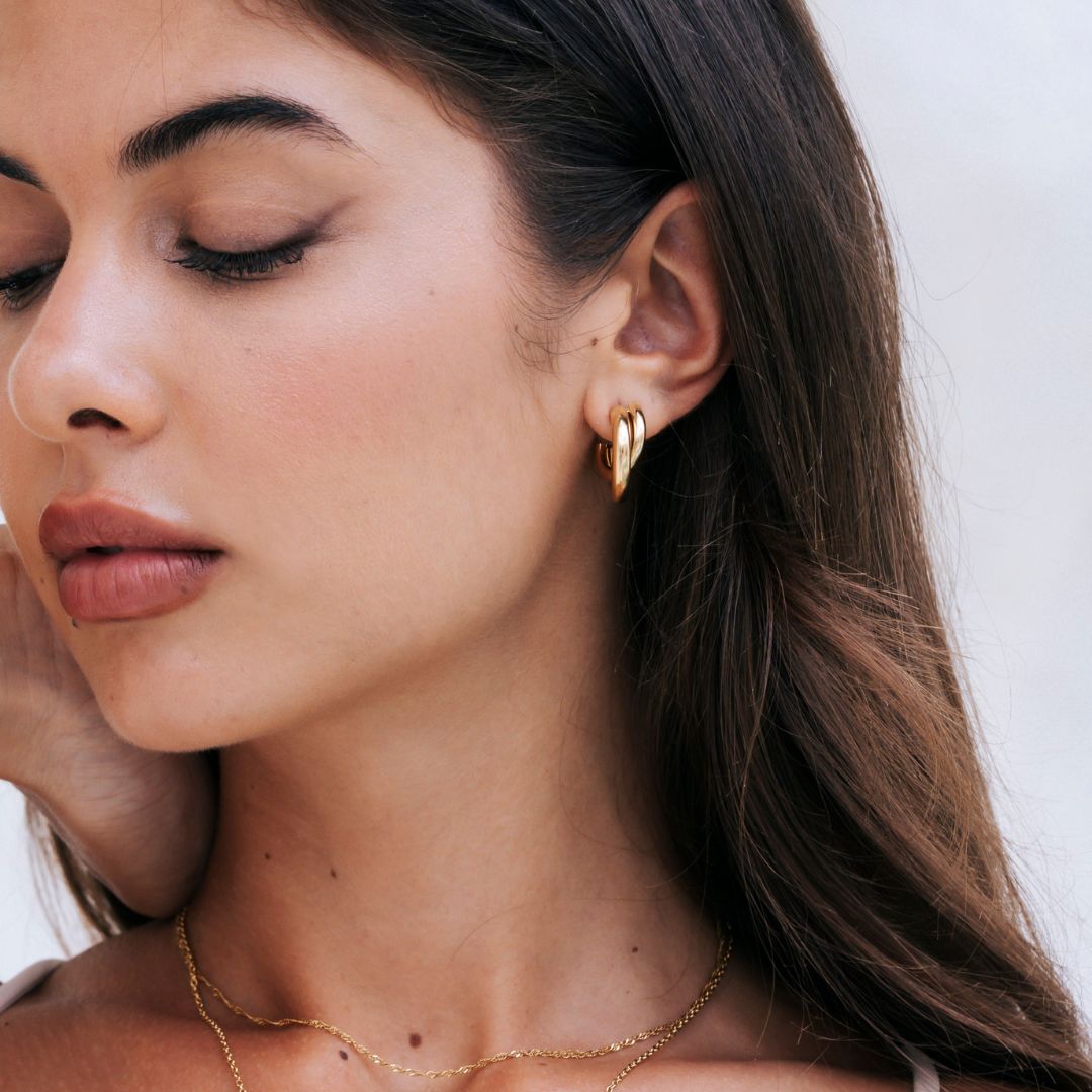 A close-up of a woman wearing the cove hoops from mettle and bloom. they are a gold hoop earring with a twist design. Her  
face partially turned away against a light neutral background.