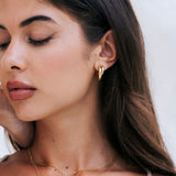 A close-up of a woman wearing the cove hoops from mettle and bloom. they are a gold hoop earring with a twist design. Her  
face partially turned away against a light neutral background.