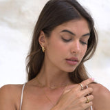A close-up lifestyle shot of a woman with her eyes closed, wearing delicate gold jewelry including a gold hoop earring, a thin gold chain necklace, and gold rings on her fingers.
