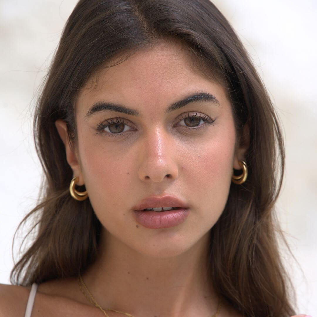 A close-up portrait of a woman with long brown hair wearing gold hoop earrings and a delicate gold necklace against a soft, light background.