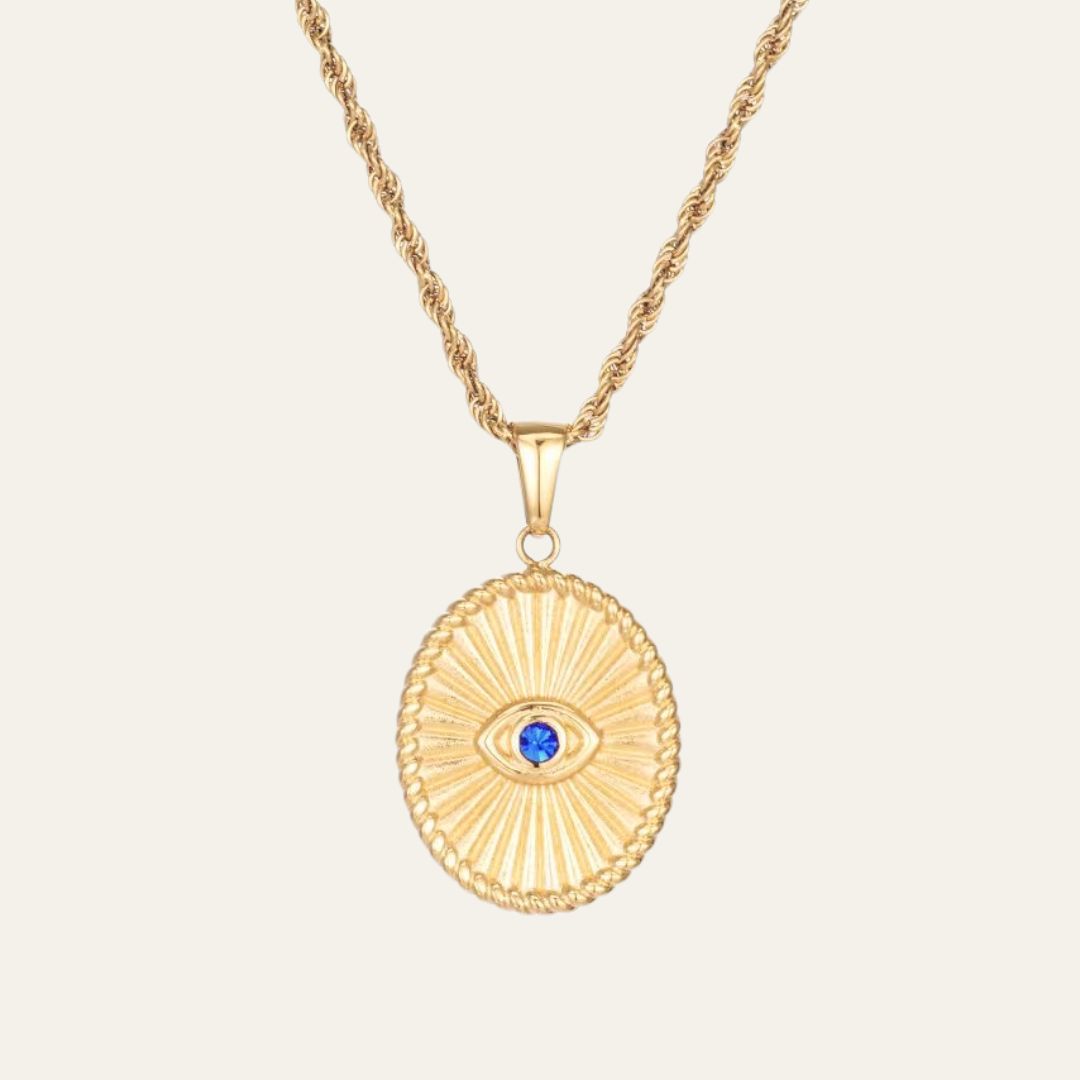 Gold necklace with a oval pendant featuring a blue gemstone on a beige background. This is the evil eye necklace. the Evil Eye wards off negativity while inviting clarity, balance, and good fortune.