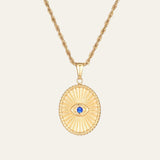 Gold necklace with a oval pendant featuring a blue gemstone on a beige background. This is the evil eye necklace. the Evil Eye wards off negativity while inviting clarity, balance, and good fortune.
