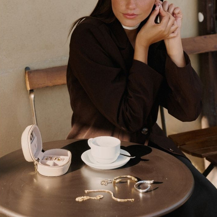 Woman sitting at a table with jewelry and a cup of coffee, in a cozy setting. She is getting ready to go out so she is taking jewellery out of her Mettle & Bloom storage case and putting the jewellery on. 