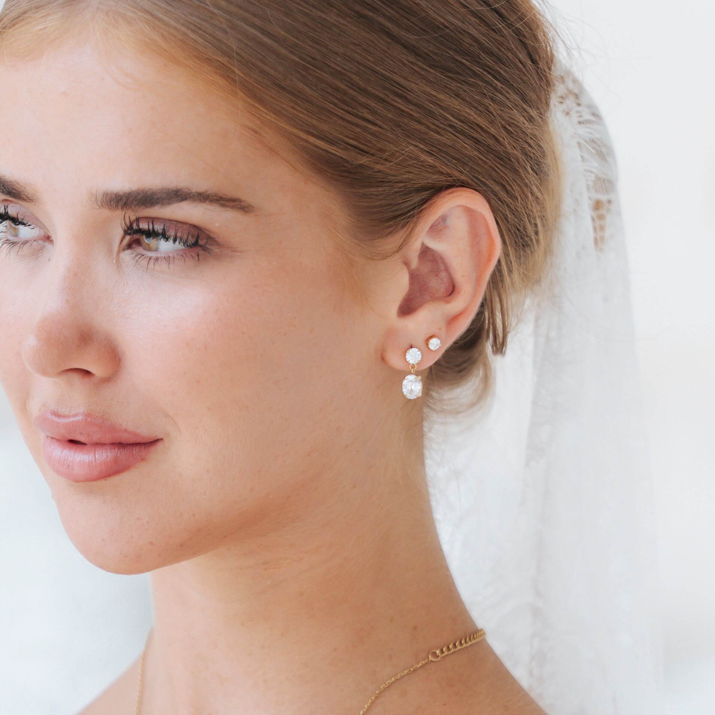 A close-up of a woman wearing delicate gold stud earrings with small white pearl-like drops, shown in profile against a soft white background with a veil.