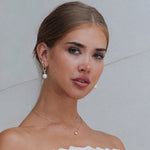 A close-up portrait of a woman wearing gold drop earrings with white pearl beads and a delicate gold necklace with a small pearl, against a light gray background.