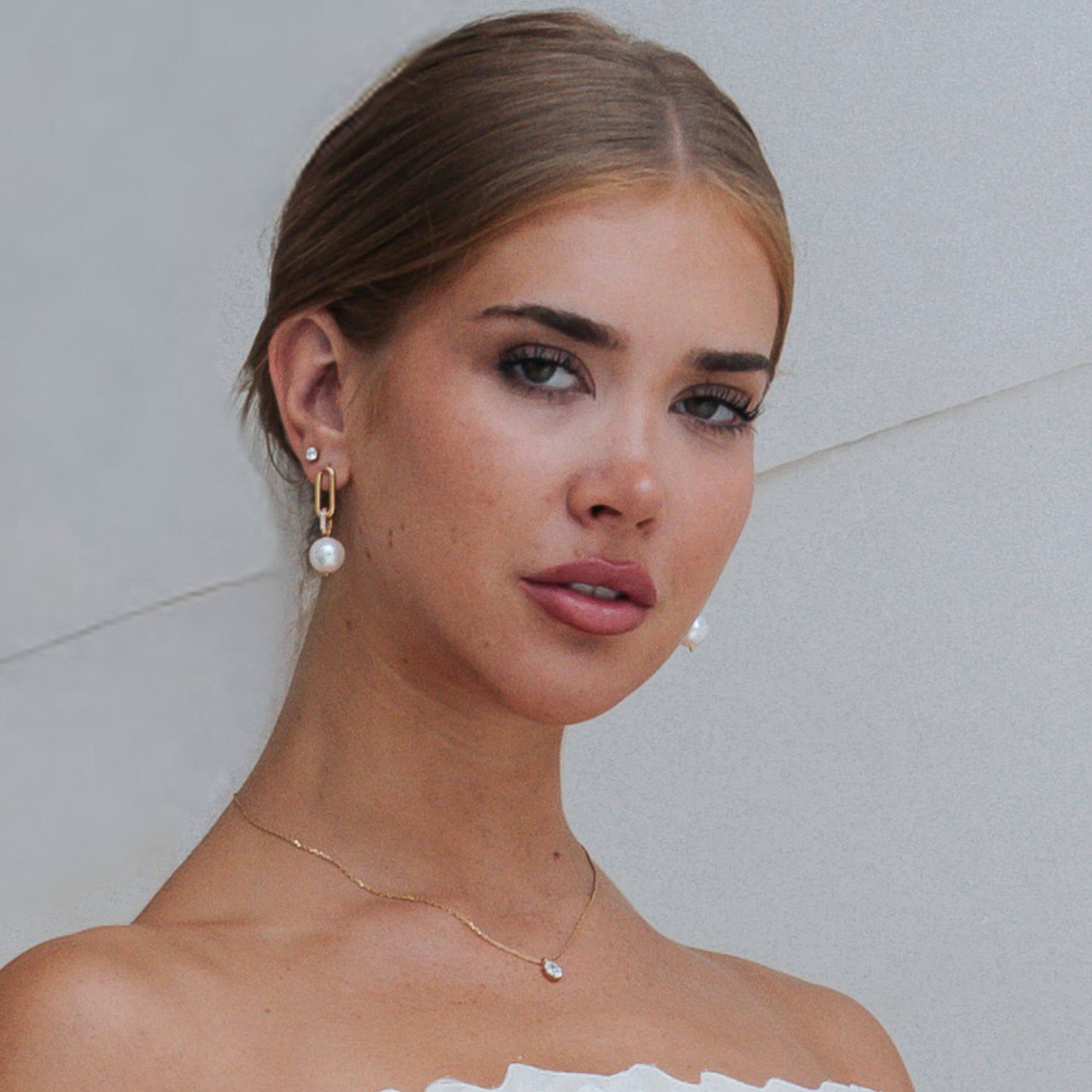 A close-up portrait of a woman wearing gold drop earrings with white pearl beads and a delicate gold necklace with a small pearl, against a light gray background.