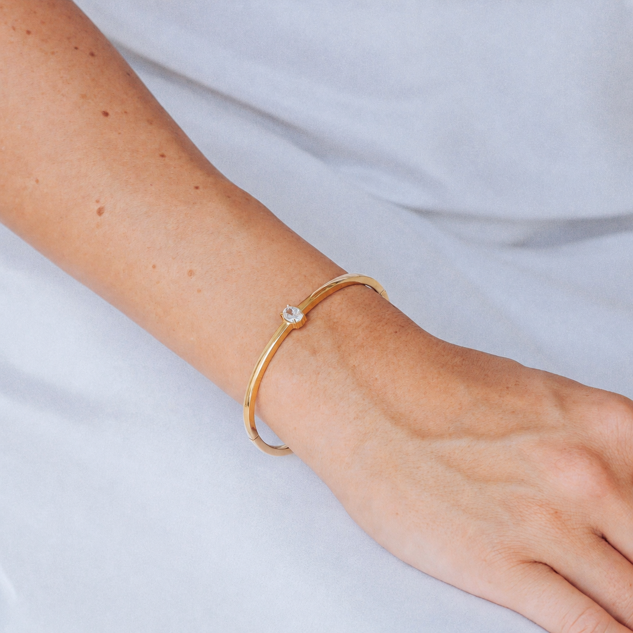 A delicate gold bangle bracelet with a single oval cut clear gemstone, worn on a person's wrist against a soft white fabric background.