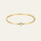 A delicate yellow gold bangle bracelet with a single rectangular-cut clear gemstone in a prong setting, shown against a solid off-white background.