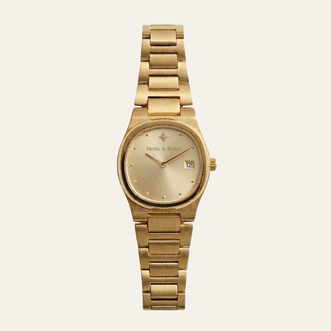 A gold-tone metal wristwatch with a matching link bracelet, featuring a champagne-gold dial with a date window and the 'Mette & Bjoern' brand name printed on the face, set against a solid off-white background.