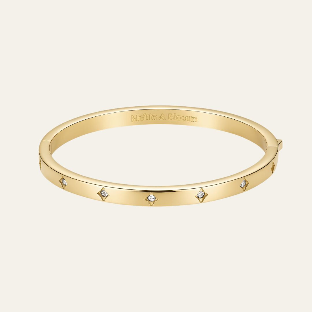 A gold bangle bracelet with small diamond accents, featuring the 'Matte & Bloom' brand name engraved inside, set against a light beige background.